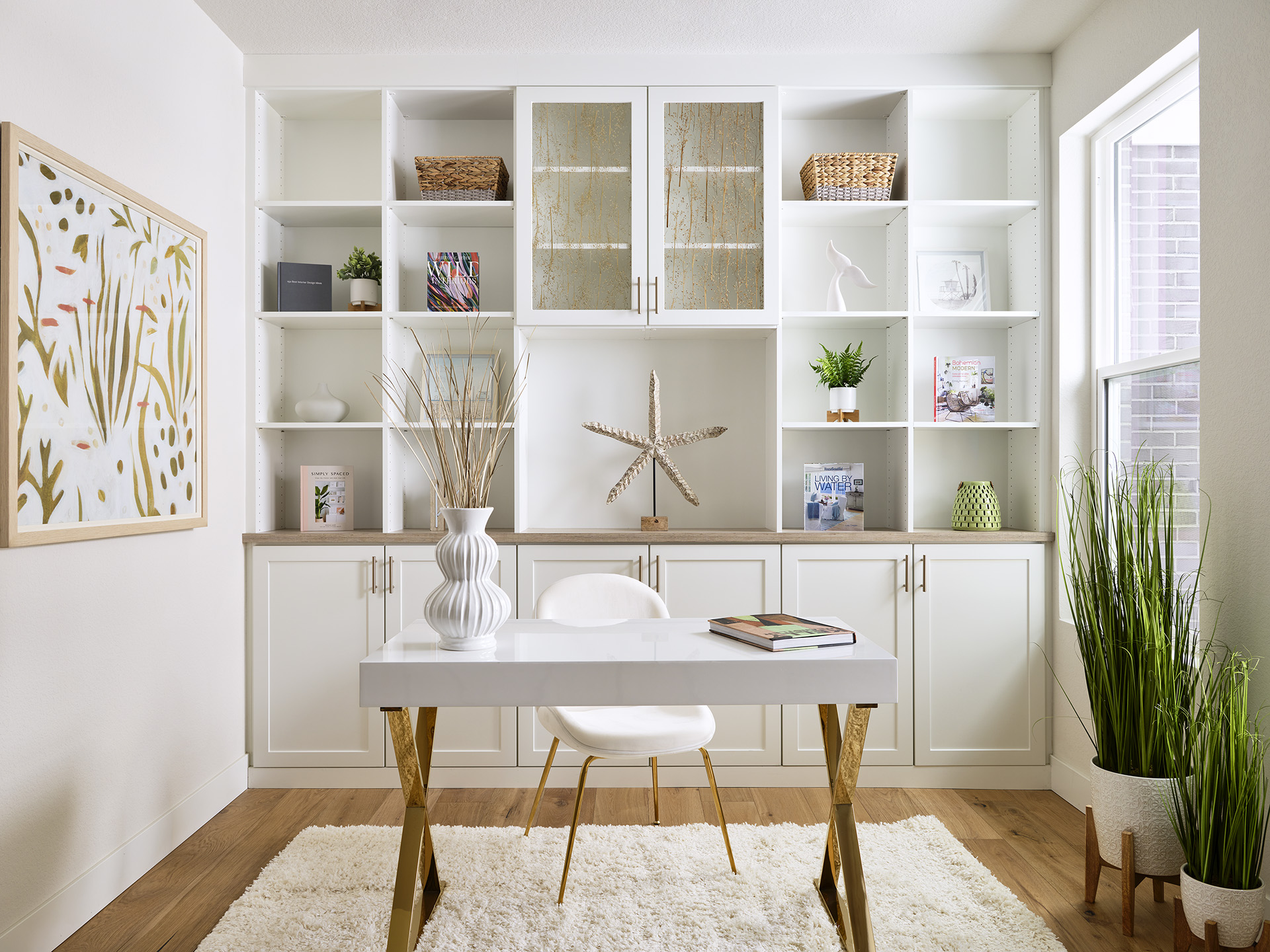A stylishly organized study with white cabinetry, desk, chair, wall art, plants, books, and decorative pieces